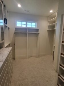 White closet shelving with hanging rods, angled shoe shelves, and a window, carpet flooring