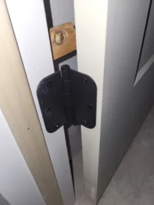 Bare wood door edge with a black hinge bracket installed and a round wood plug set nearby
