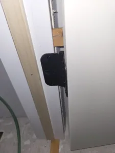 Door edge with a black bracket screwed on and a wood shim wedged into the jamb gap