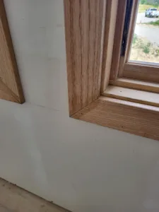 Wood window jamb corner with a mitered joint, unfinished, drywall visible, floor below