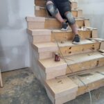 Worker finishing raw wood stair treads and risers with stain or sealant during trim stage.