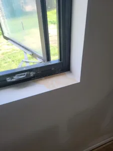 Primed window casing installed at an interior corner, black framed window with grass visible outside.