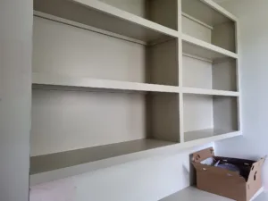 Built-in open shelving with two main shelves and corner shelf, painted, cardboard box stored below.