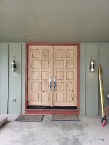Natural wood double exterior doors with raised panels, flanked by wall sconces at entry.