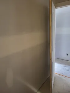 Wall filling the foreground in dim light, with a white door open at right into a room with bare subfloor.