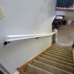 Painted white wall-mount handrail with brackets installed on stairwell wall alongside carpeted stairs.