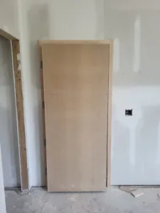 Unfinished raw wood door slab standing upright in a finished drywall room, awaiting installation.