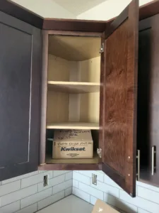 Open stained cabinet door showing interior shelves and a boxed lock set, near tile backsplash