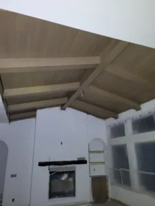 Wood plank vaulted ceiling with parallel beams over an unfinished room with plastic covered windows