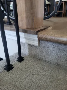 Wood nosing cap wrapping a stair newel post base where flooring meets carpeted treads