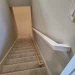Wood handrail mounted to stairwell wall with white painted casing and carpet treads installed.