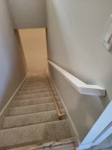 White handrail mounted to the wall above a straight staircase with carpet treads and white trim.