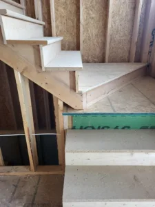 Primed staircase treads and risers under construction within open stud wall framing, OSB subfloor visible.