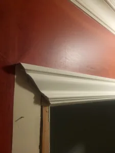 White painted crown molding wraps a wall corner above a doorway, dark red wall and raw framing visible.