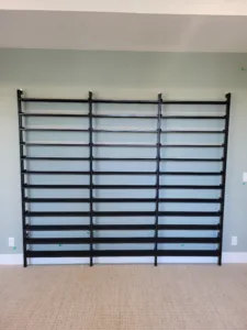 Black metal shelving rack with multiple horizontal rods installed on a wall in an empty carpeted room.