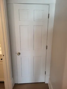 Painted white six-panel interior door closed, casing and hinges installed, over wood-look flooring.