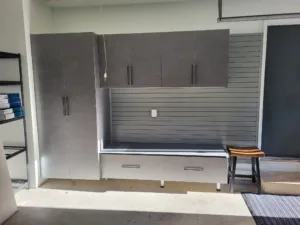 Garage cabinet wall with gray upper cabinets, center slatwall panel, and lower storage bench, fully installed.