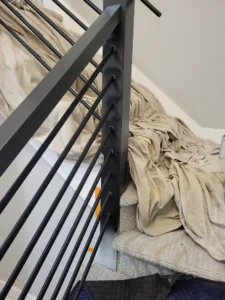Black metal stair balusters fastened into a diagonal rail post, treads covered during finish work