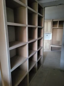 Tall unfinished cubby bookcase built into wall opening onto hallway with cabinetry, raw wood visible.