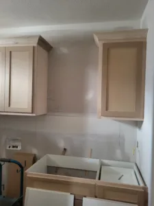 Kitchen cabinet install with unfinished wood upper cabinets and lower boxes, drawers removed, ready for paint.