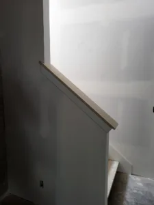 Wood cap on a painted charcoal knee wall at a stairway opening, with bare drywall beyond.
