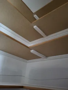 Coffered ceiling corner where white crown molding trims raw wood panel beams and framing.