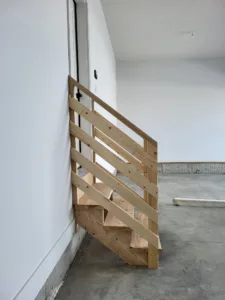 Unfinished wood garage steps with horizontal slat railing under construction against interior wall.