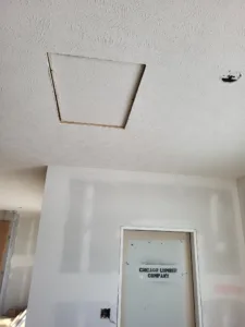 Attic access opening cut into textured ceiling with rough edges, hallway below has taped drywall walls