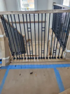 Closeup of vertical black metal balusters installed below a wood cap rail, taped subfloor visible
