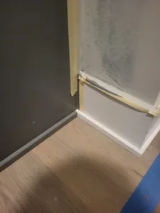 Painted white baseboard return taped at inside corner, wood look flooring visible below.