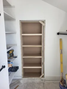 Unfinished built-in shelving with five shelves in a boxed wall recess, tools left on the floor.