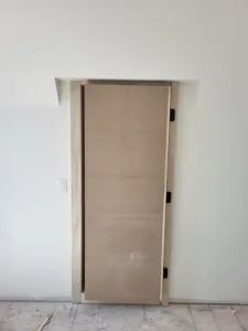 Door slab hung with black hinges in unfinished opening, raw wood casing not yet painted.