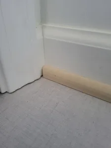 Natural wood baseboard meets wall at floor line, white casing visible above, unfinished stage.