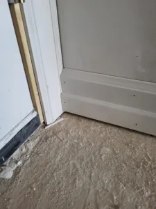 Painted white baseboard meeting a door jamb over a dusty subfloor, with a dark door sweep nearby