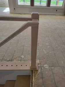 Raw unfinished wood newel post and angled handrail piece resting on plywood subfloor
