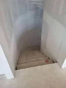 Staircase descending through a narrow stairwell with curved drywall walls ready for paint.