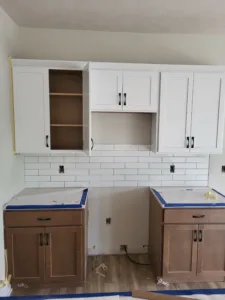 Kitchen mid-install with painted white upper cabinets, subway tile backsplash, and wood lower cabinets.