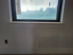 Raw wood window sill installed below a black framed window, unpainted, overlooking a tree line outside.