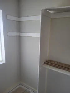 White closet shelves meeting at an inside corner with a wood hanging rod below, natural window light.