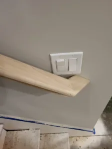 Unfinished wood handrail with a curved return end, mounted near a wall light switch
