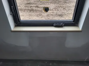 Raw wood sill with white casing legs at a black framed window, dirt lot visible outside.