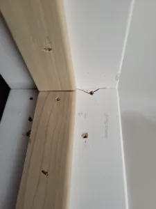 Mitered white trim corner joint with drilled screw holes, natural wood rail behind, mid install