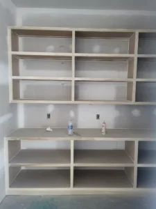 Unfinished built-in cubby shelving with a mid-height counter shelf and electrical outlets, mudroom style.