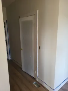 Primed grey interior door with brass knob, blue tape along casing near the floor, wood-look flooring.