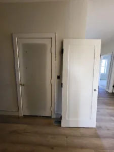 Old flush door hung in its frame beside a new white panel door slab leaning nearby in a finished hallway.