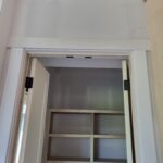 Closet opening with painted jamb, casing, and black hinges installed; shelving interior visible, doors not yet hung.
