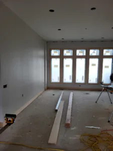 Baseboard stock staged on the subfloor in a room with finished drywall and windows, before install.