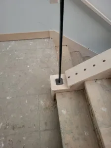 Black round post base mounted on a wood platform near a raw stair stringer piece, unfinished