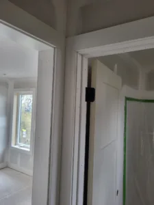 Painted white door hung on a black hinge in a finished frame, with a window visible down the hall.