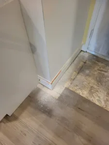 Painted white baseboard installed along a corner where wood-look flooring meets bare subfloor.